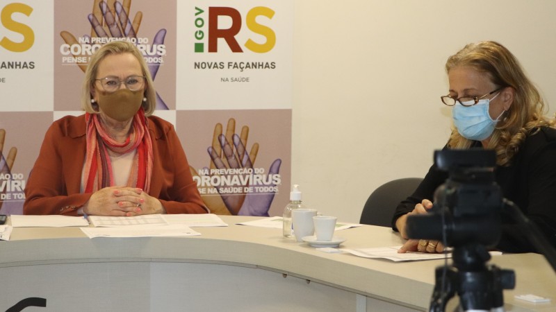 Secretária Arita olha para a frente enquanto Tani olha para um papel que está na mesa. As duas estão sentadas, de máscara.