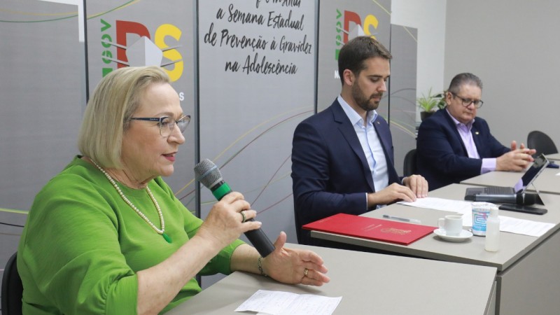 Secretária Arita fala, segurando o microfone. Governador Leite e secretário da Segurança Pública, Ranolfo Vieira Júnior, observam. Todos estão sentados atrás de uma mesa.