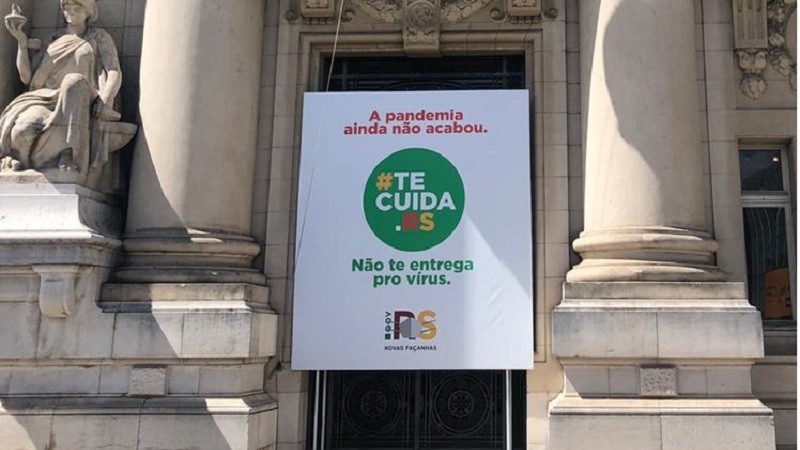 Imagem do banner da campanha Tecuida.RS no Palácio Piratini. No banner, está escrito "A pandemia ainda não acabou. Não te entrega pro vírus". 
  