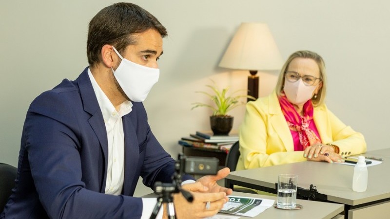 Governador Leite olha para frente e fala enquanto a secretária Arita, à sua direita, a observa. Os dois estão sentados atrás de uma mesa e de máscara.