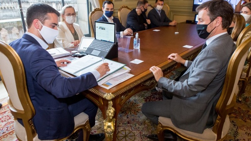 Governador Leite faz anotações enquanto participantes da reunião o observam. Todos estão de máscara e sentados em volta de uma mesa.