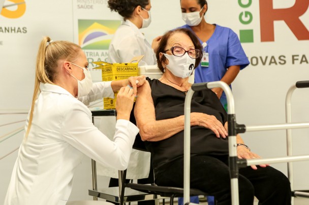 Dona Eloina, sentada e de óculos, recebe a vacina dada por uma profissional de saúde. As duas estão de máscara. 