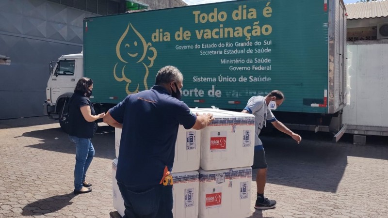 Dois funcionários, de máscaras, empurram lotes de vacinas em direção ao caminhão. Ao lado, uma outra funcionária, também de máscara, acompanha.  