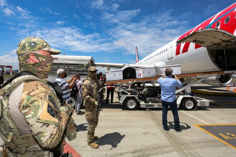 A imagem mostra um lote de vacinas entrando no avião enquanto alguns homens fotografam e outros fazem a segurança.