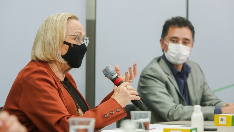 A secretária Arita fala, segurando um microfone. Bruno Naundorf está a seu lado. Os dois estão sentados, de máscara.  