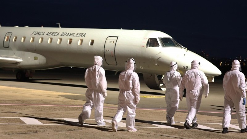 A imagem mostra cinco profissionais de saúde uniformizados caminhando, num primeiro plano, e uma aeronave ao fundo. 