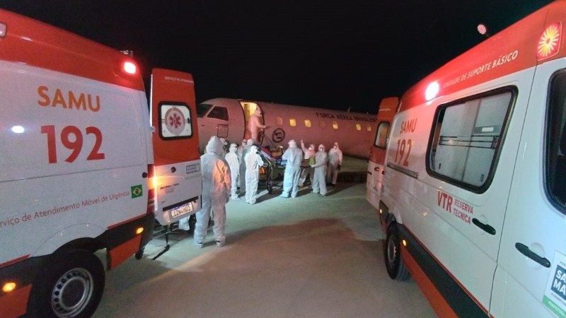 A imagem mostra duas ambulâncias em primeiro plano e profissionais de saúde uniformizados embarcam um paciente acamado numa aeronave. 