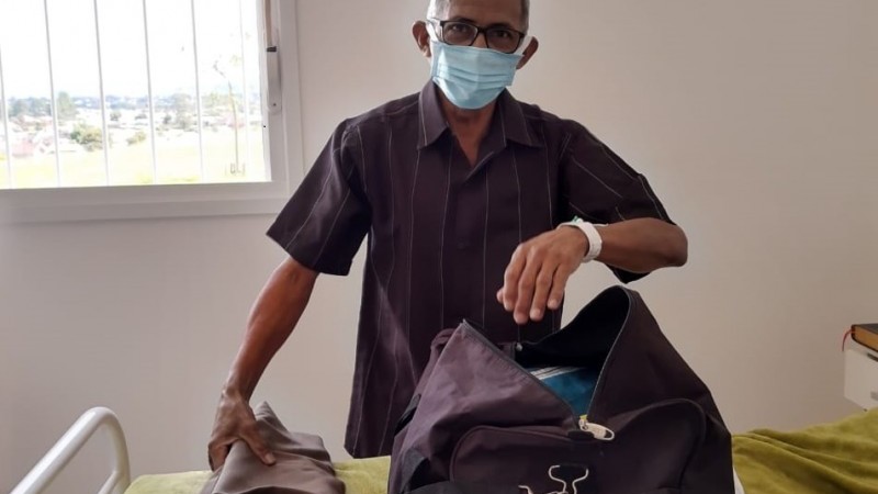 João, de máscara, acomoda suas roupas na mala, que está em cima de uma cama.