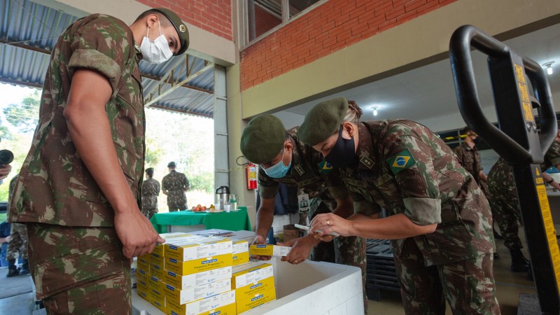 Três militares, de máscara, observam e manuseiam caixas de medicamentos, que estão numa caixa grande de isopor.