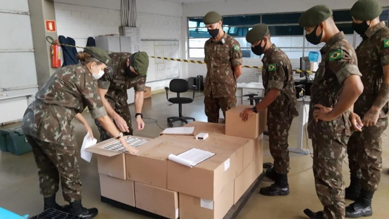 A imagem mostra dois militares mexendo em caixas com vacinas. Outros quatro observam. Todos est&atilde;o de m&aacute;scara. 