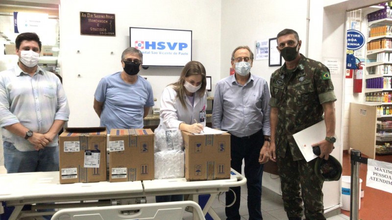 Uma profissional de saúde, um militar e mais três homens, de pé e de máscara estão num depósito cheia de caixas e prateleiras.  