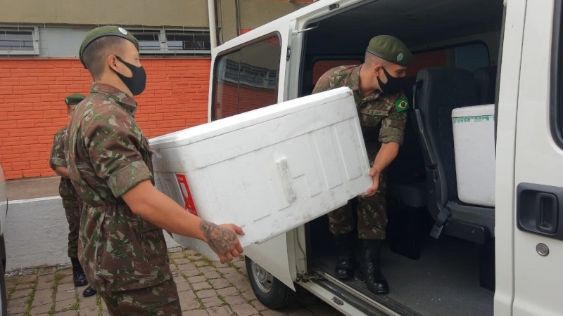 Dois militares, de máscara, colocam uma caixa grande de isopor dentro de uma van. 