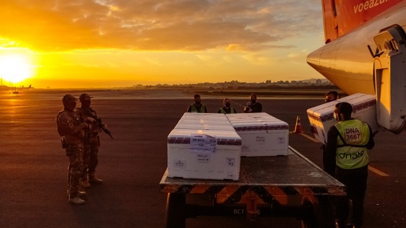 A imagem mostra um reboque com várias caixas com doses de vacinas, dois funcionários tirando outra caixa do avião para botar no reboque. Alguns militares e funcionários observam a cena, que acontece num final de tarde.