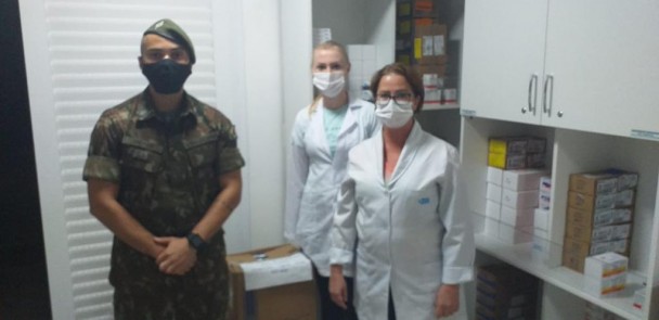 Um militar e duas profissionais de saúde, de pé e de máscara, olham para a frente numa sala com armários e caixas de remédios. 