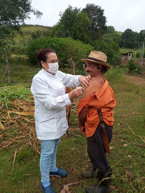 Uma profissional de saúde vacina um homem de chapéu no meio do campo. 