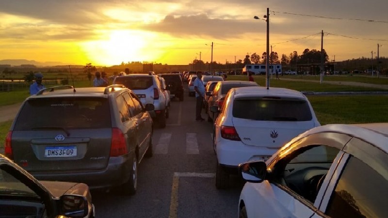 De manhã, com o nascer do sol, vários carros e algumas pessoas esperam a vacinação no asfalto.  