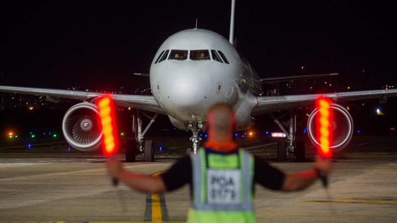 A imagem mostra, ao fundo, um avião de frente. Em destaque, um funcionário com sinalizadores luminosos.  