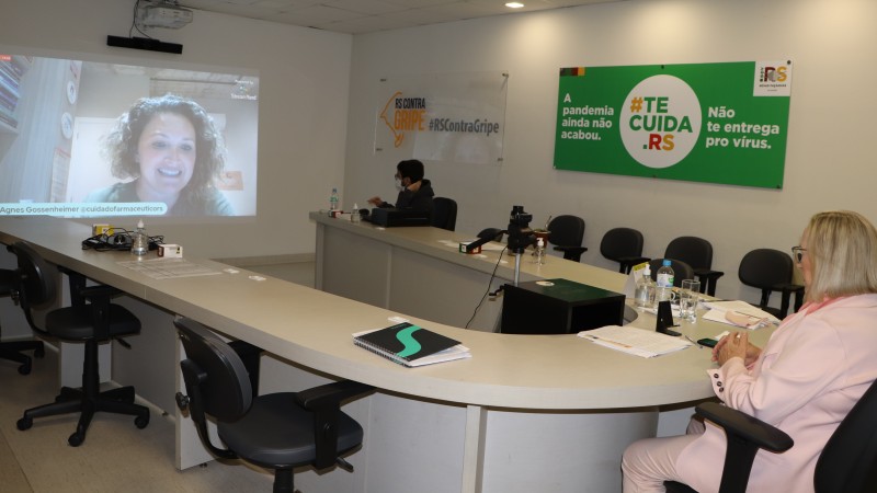 Secretária Arita, sentada atrás de uma mesa, assiste a fala de participante da live que está sendo projetada no telão. 
