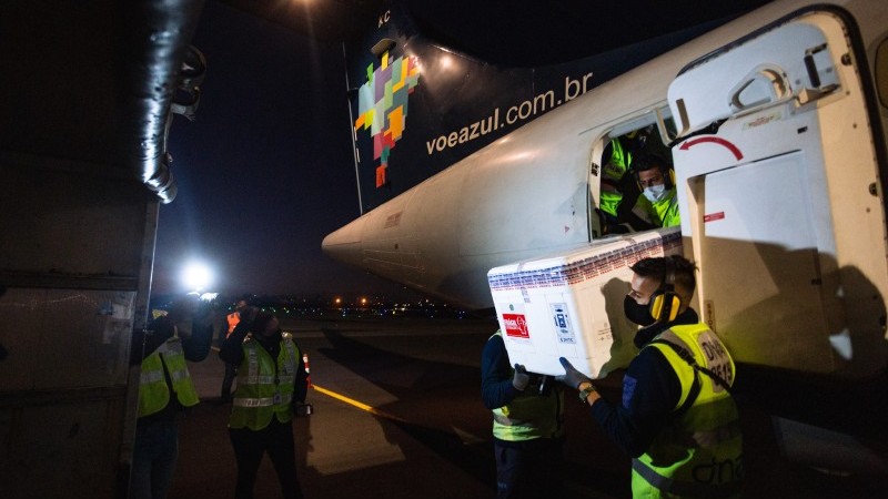Alguns funcionários seguram uma caixa grande com várias doses de vacinas em torno de um avião. Outros funcionários observam a cena, que acontece à noite.  