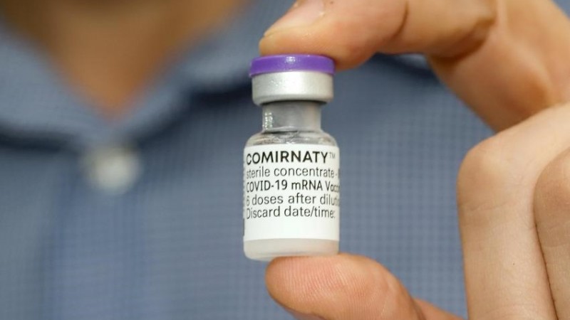 A imagem mostra uma mão segurando um frasco da vacina. 