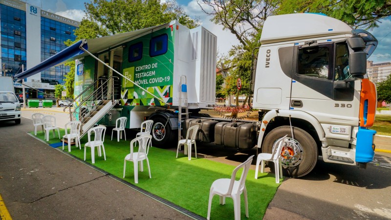 A imagem mostra um trailer do Centro de Testagem Móvel para a Covid com algumas cadeiras de plástico em cima de um tapete que está na frente do trailer.
