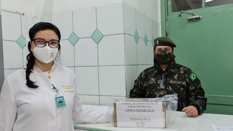 Militar e profissional de saúde, de máscara, olham para a frente e posam com uma caixa que está em cima de um balcão. 