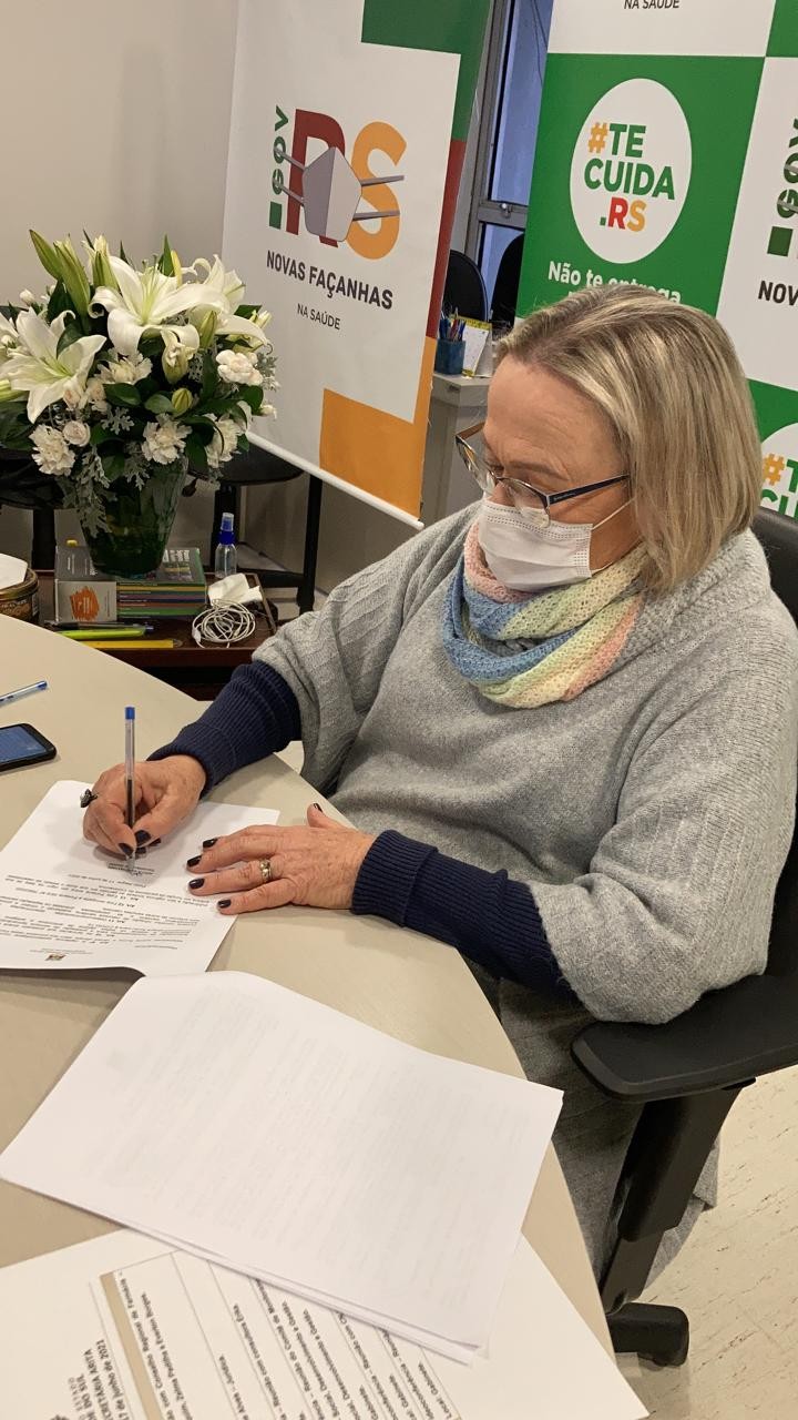 Secretária Arita, sentada e de máscara, assina um documento.