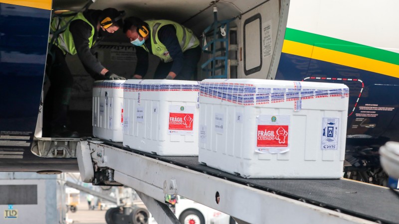 Dois funcionários ajeitam caixas grandes com doses de vacinas na esteira.