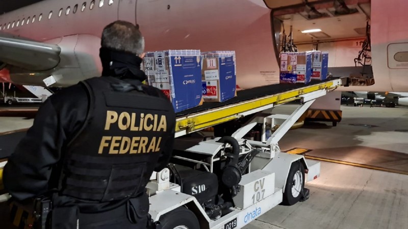 Funcionário da Polícia Federal observa duas caixas grandes com doses de vacinas deslizando pela esteira. 