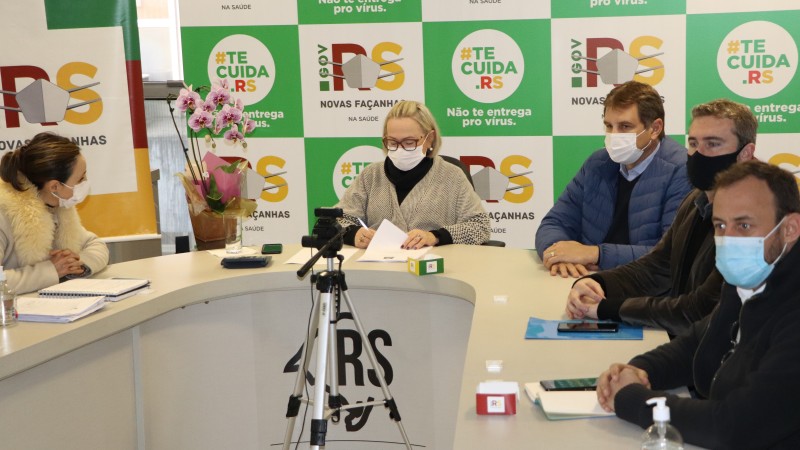 Secretária Arita assina um documento enquanto outros quatro participantes da reunião acompanham. Todos estão de máscara e sentados atrás de uma mesa. 