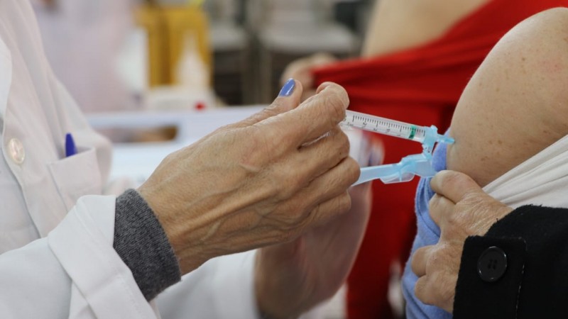 Imagem mostra uma mão de uma profissional com a seringa aplicando a vacina em uma mulher em uma Unidade de Saúde.