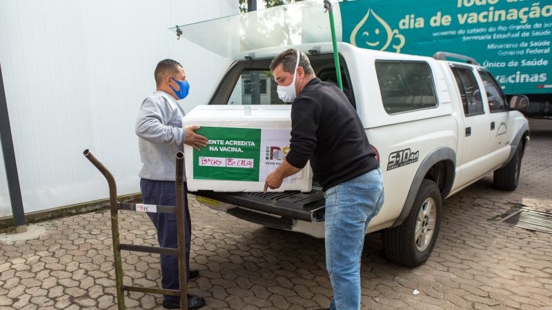 Dois funcionários, de máscara, tiram uma caixa grande de isopor com doses de vacinas da parte traseira da caminhonete.
