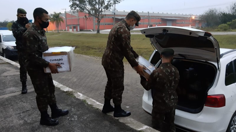 Três militares carregam caixas para o porta malas de um carro branco.