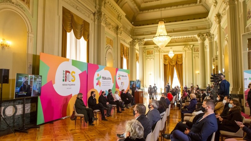 Imagem panorâmica do evento com participantes e público, sentados um de frente para o outro. 