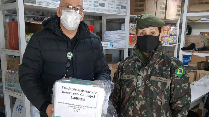 Um homem segurando uma caixa com as inscrições da Fundação de Camaquã , ao lado de uma militar do exército fardada, os dois usam máscaras de proteção contra o coronavírus. Os dois estão num depósito. 