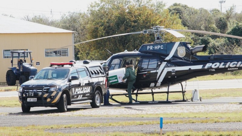 Dois homens carregando uma caixa para dentro do helicóptero. Na frente, uma caminhonete da Polícia.