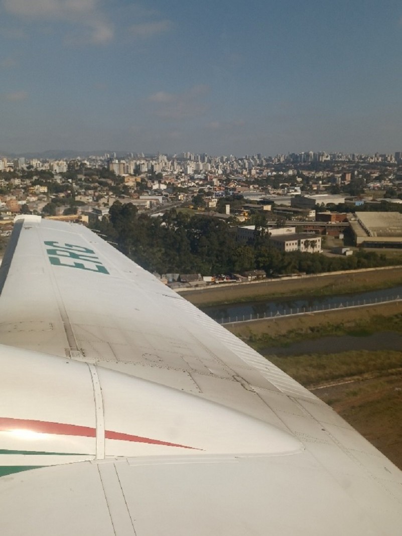 Imagem aérea feita da janela da avião mostra uma cidade com seus prédios, casas, fábricas e árvores.