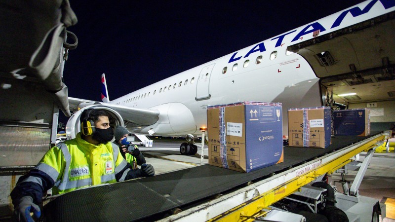 Um funcionário, de máscara, acompanha as caixas que descem pela esteira que sai do avião. 