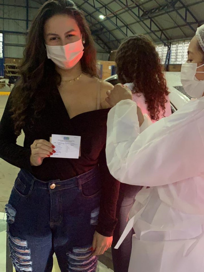 Tatiany, sorrindo e segurando a carteira vacinal, recebe a vacina de uma profissional de saúde. As duas estão de pé e de máscara.  