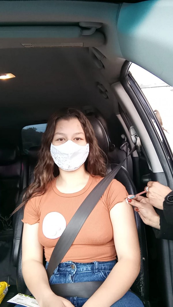 Sentada dentro do carro, Gabriela, de máscara, recebe a vacina de uma profissional de saúde. 