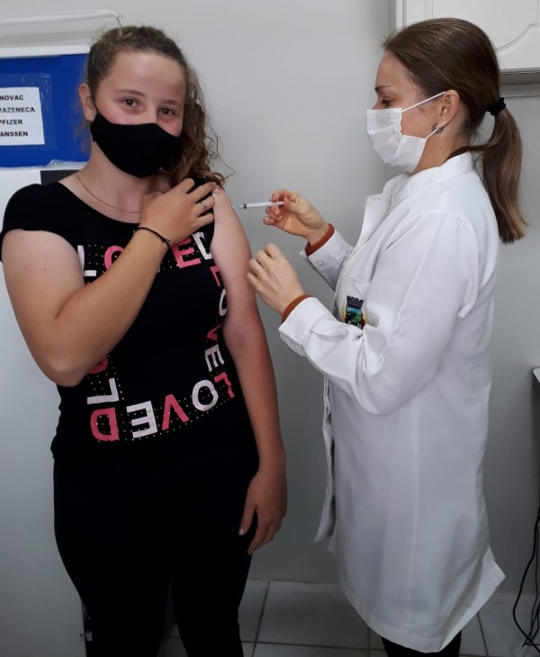 Gyovanna recebe a vacina de uma profissional de saúde. As duas estão de pé e de máscara.