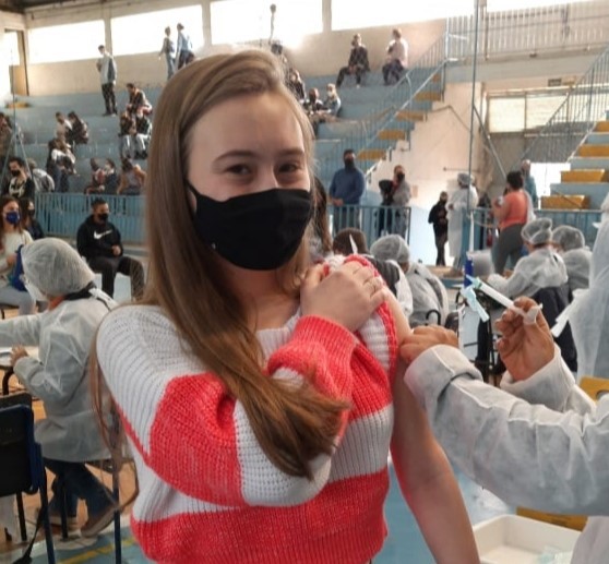 A estudante Helena Aurélio Temp, de 19 anos, recebe a vacina de uma profissional de saúde. As duas estão de pé e de máscara.