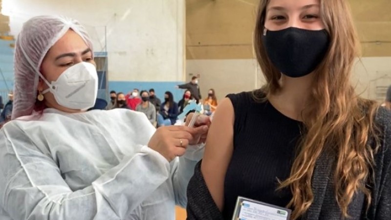 A estudante de arquitetura Fernanda Escarrone Bonaza segura a carteira vacinal enquanto recebe a vacina de uma profissional de saúde. As duas estão de pé e de máscara.