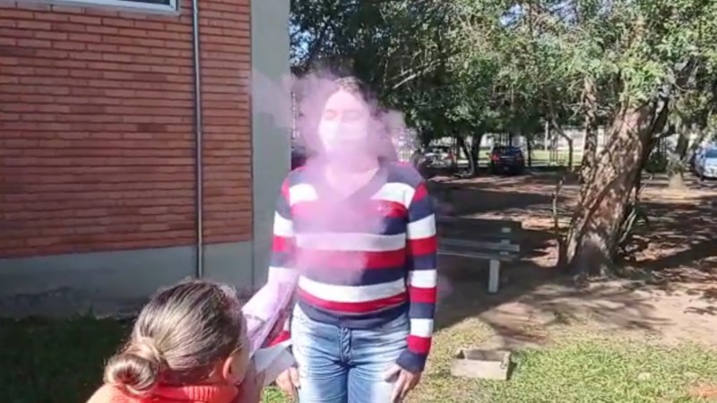 Uma mulher abaixada assopra fumaça colorida atóxica para outra mulher que está de pé e de máscara. A cena ocorre ao ar livre, num gramado.