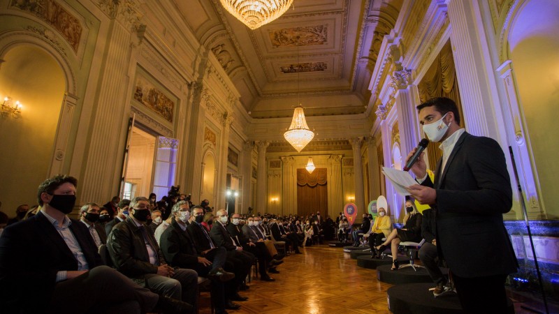 De pé e de máscara, Governador Leite fala segurando o microfone enquanto plateia assiste sentada. 