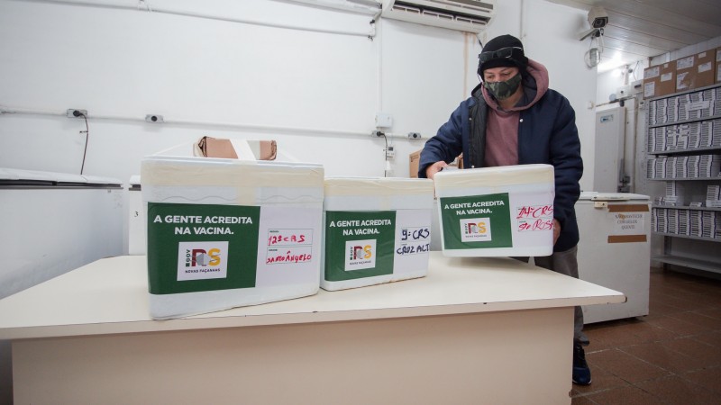Um funcionário, de máscara, coloca uma caixa de isopor com vacinas em cima de uma mesa grande ao lado de outras duas caixas.