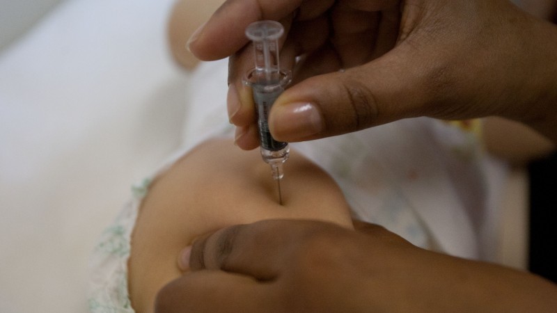 A imagem mostra duas mãos dando uma vacina na perna de um bebê. 