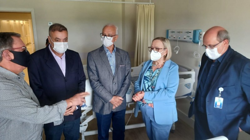 Secretária Arita ouve um dos participantes da visita. Outros três homens também ouvem. Todos estão pé e de máscara. A conversa acontece num quarto de hospital, perto de um leito equipado.