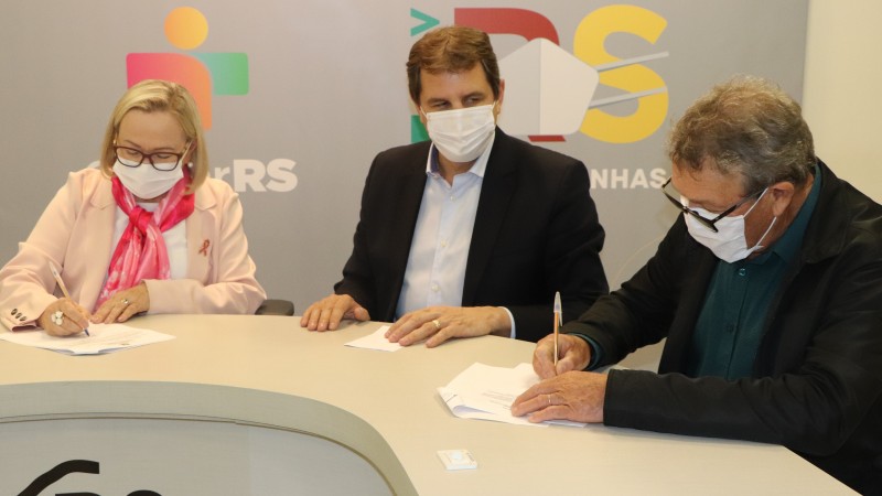 Secretária Arita e Davi assinam um documento. Outro participante da reunião assiste o momento. Os três estão sentados atrás de uma mesa e de máscara. 