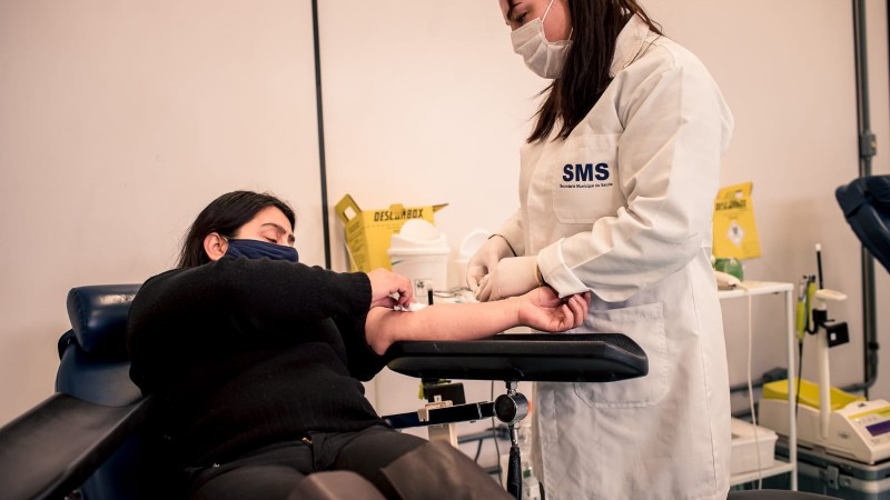 Uma profissional de saúde tira sangue de uma doadora que está sentada na cadeira.  